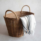 Brown wicker tote basket with two handles holding a throw