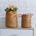 Two tall natural woven baskets with leather handles, one with flowers