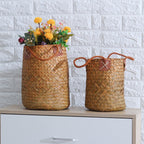 Two tall natural woven baskets with leather handles, one with flowers