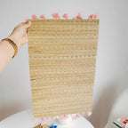 Rectangular hand woven straw placemat with pink cotton tassels at the edges